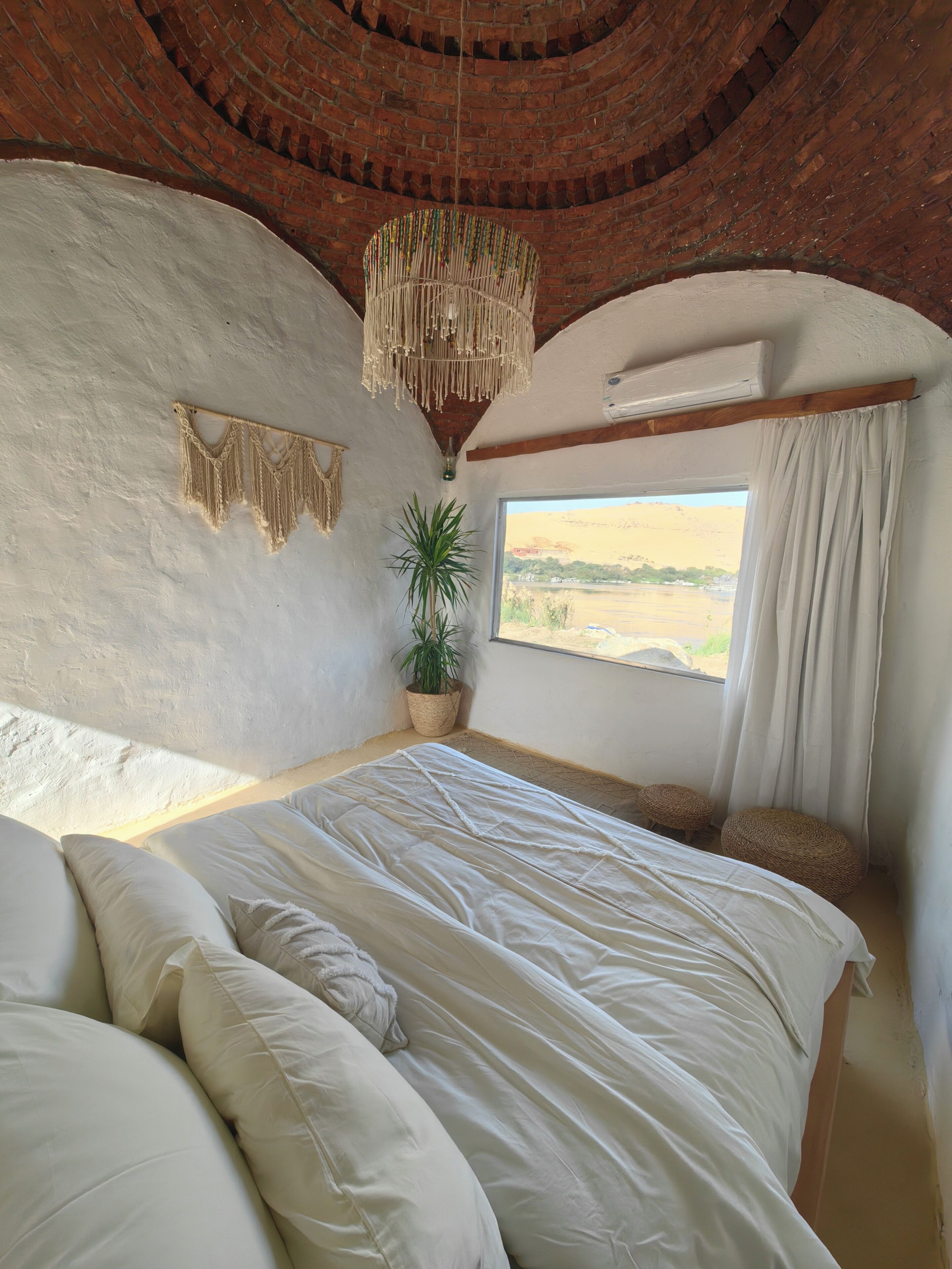 Un dormitorio con techo de ladrillo, cama con ropa de cama blanca, lámpara colgante de macramé, planta en maceta y vista de un paisaje a través de una ventana.