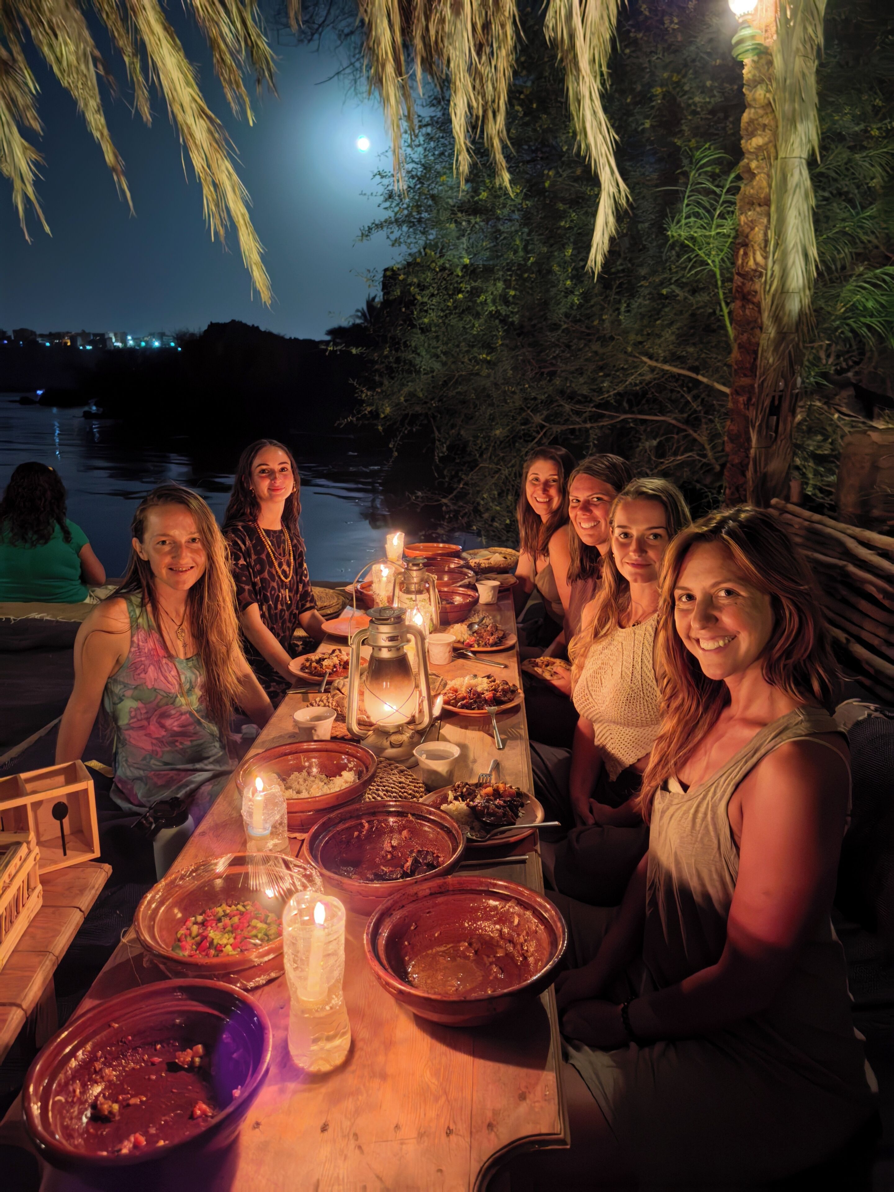 Un grupo de personas cenando al aire libre de noche, con una mesa dispuesta con comida y velas, bajo un árbol con ramas colgantes.