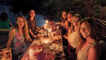 Un grupo de personas cenando al aire libre de noche, con una mesa dispuesta con comida y velas, bajo un árbol con ramas colgantes.