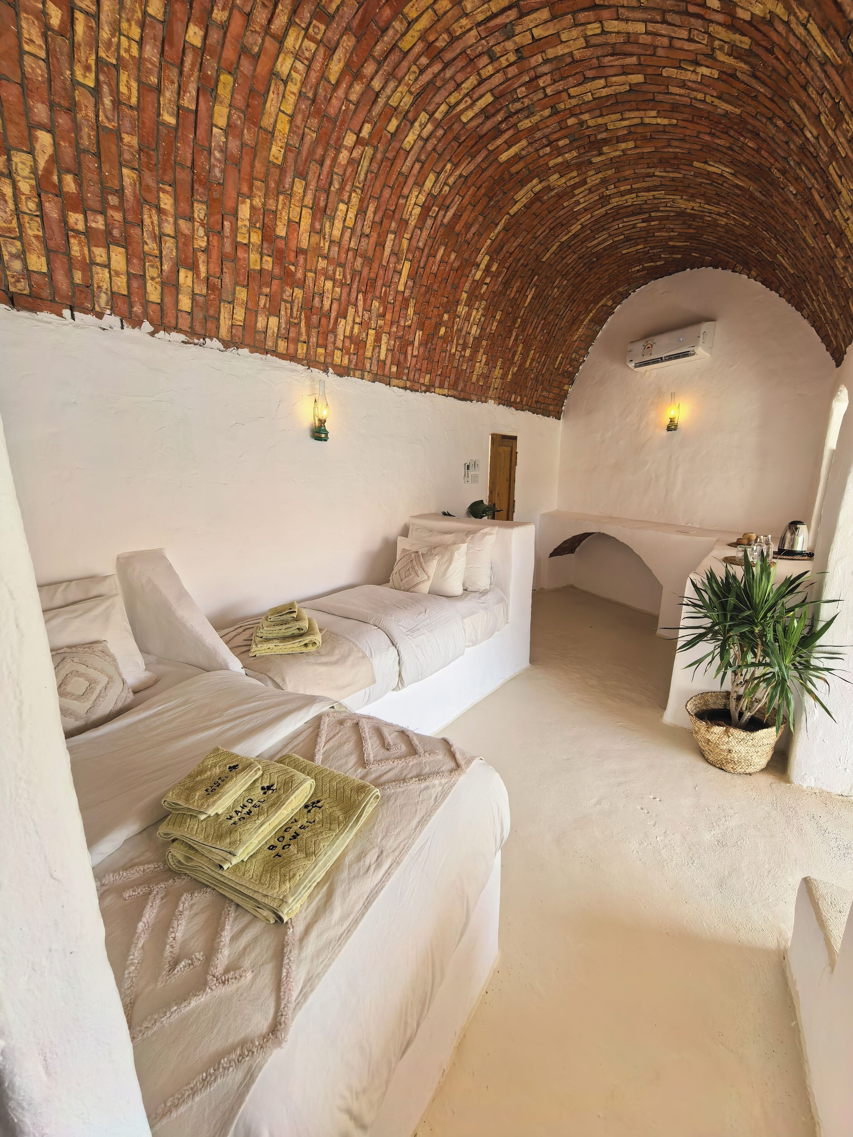 A room with a brick arched ceiling, two beds with white bedding, a small table, a potted plant, and a wall-mounted air conditioner.