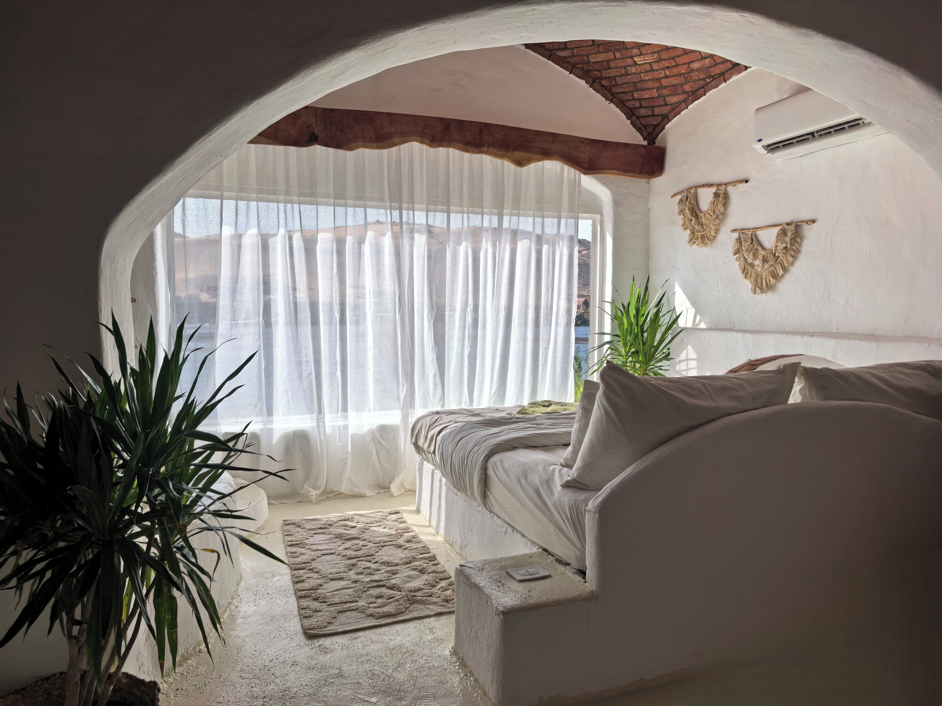 A bedroom with a bed, a window with sheer curtains, a plant, and a view of the outside.