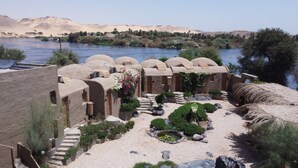A desert resort with thatched-roof buildings, a river, and a mountainous backdrop. - Nala Narty Nubian house and organic farm (Aswan)