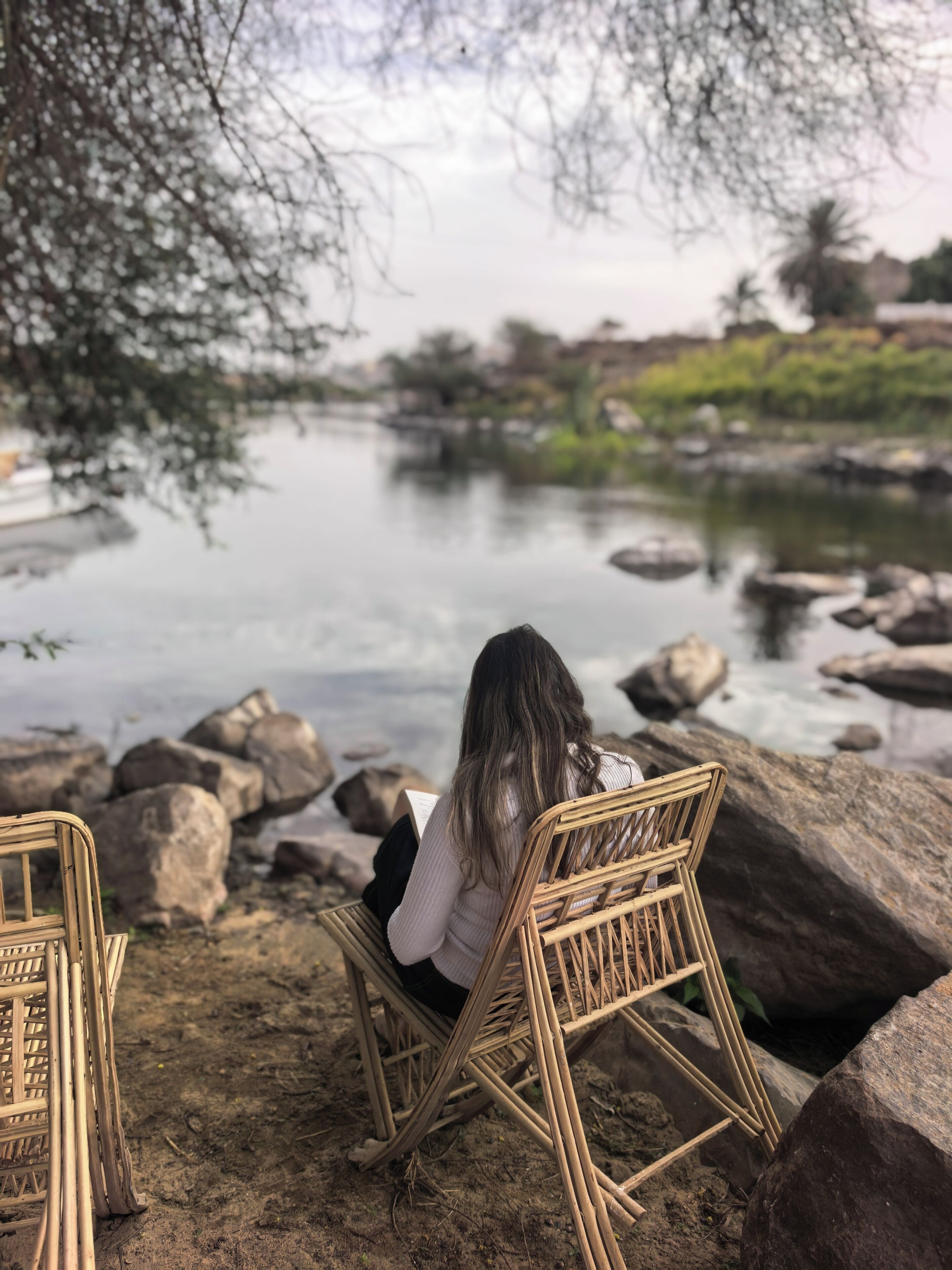 A person sitting on a wicker chair by a river, with rocks and greenery in the background.