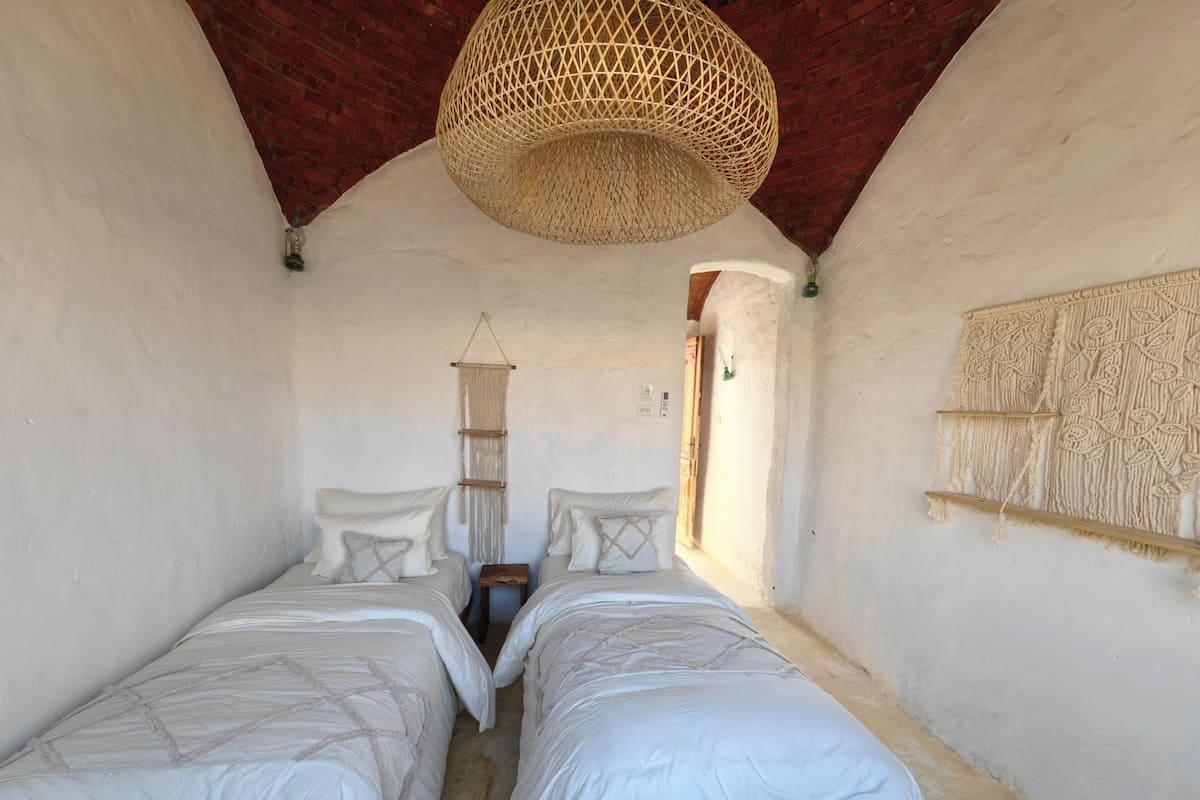 Une pièce avec deux lits, une décoration murale en macramé, un luminaire en fibres tressées et une porte en bois.
