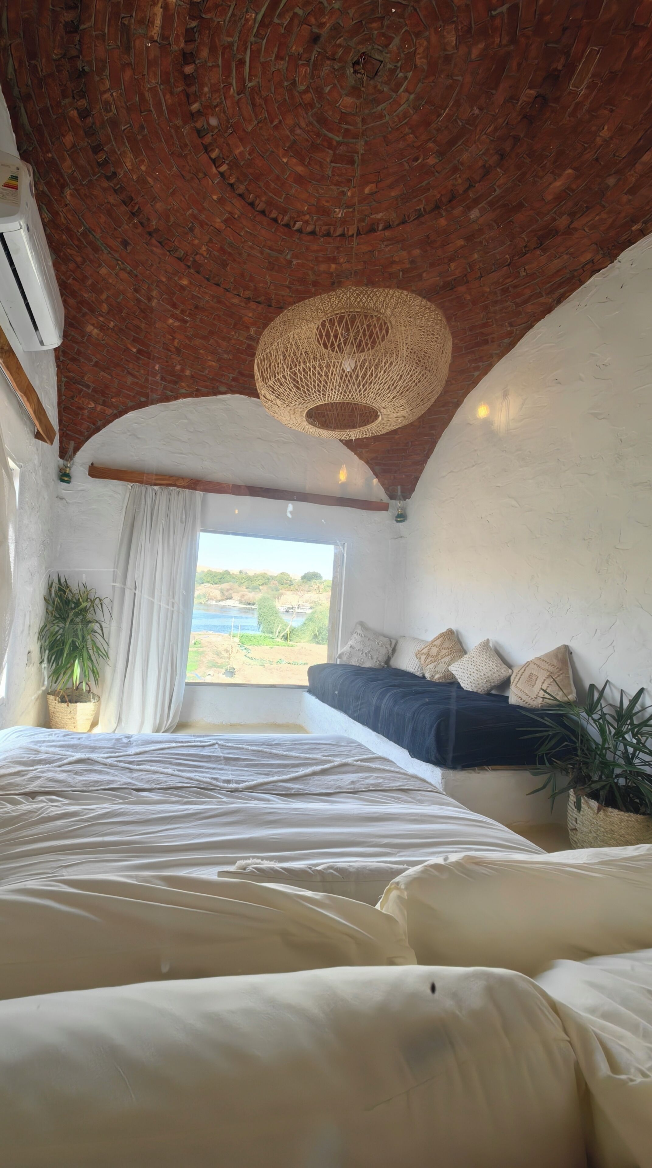 Ein Schlafzimmer mit Backstein-Decke, einer Hängeleuchte aus Rattan, einem Bett mit weißen Bettwäsche, einem Fenster mit Blick ins Freie und einem Wandregal mit Kissen.