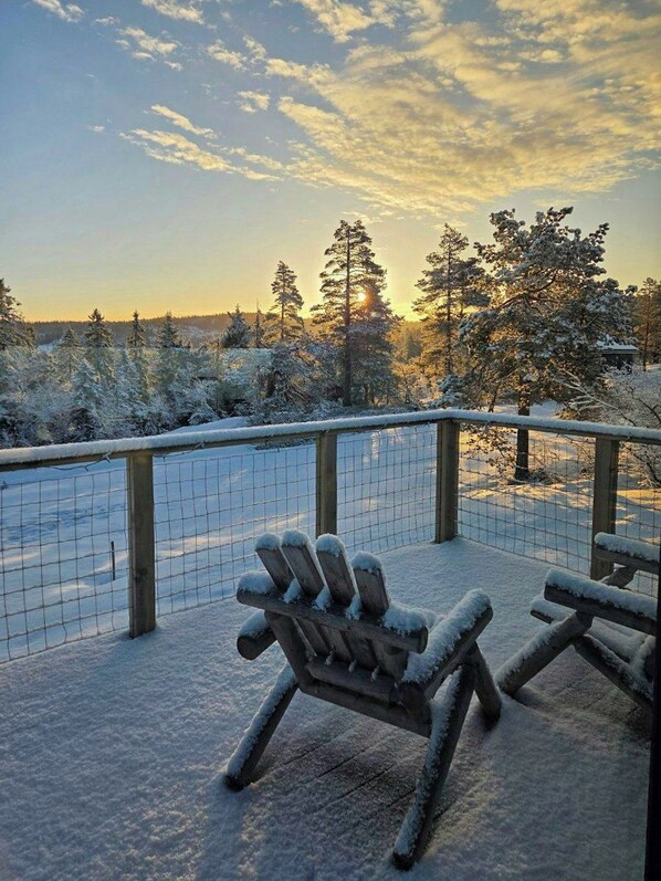 A snow-covered deck with two Adirondack chairs, a wooden fence, and a view of snow-laden trees under a partly cloudy sky.