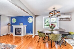 A dining area with a fireplace, a round table with chairs, and a window with a view of trees. - Corn Hill Ranch - East Falmouth (Falmouth)