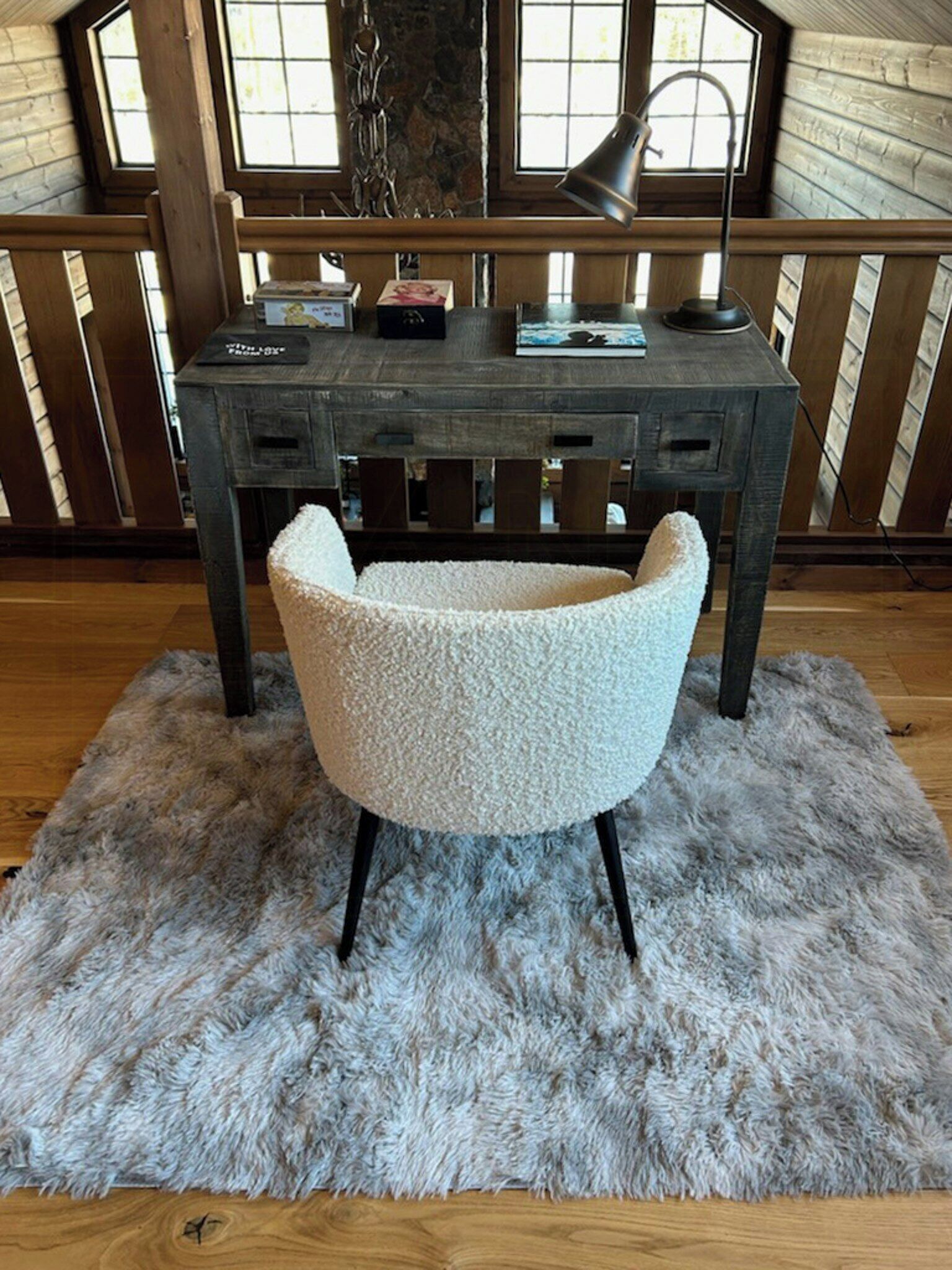 A rustic wooden desk with a lamp, a plush chair, and a shaggy rug in a room with wooden walls and large windows.