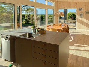 A modern kitchen with a built-in dishwasher, wooden cabinets, a large window, and a dining area with a table and chairs. - Beautiful setting by the sea with light and sky (Grenå)