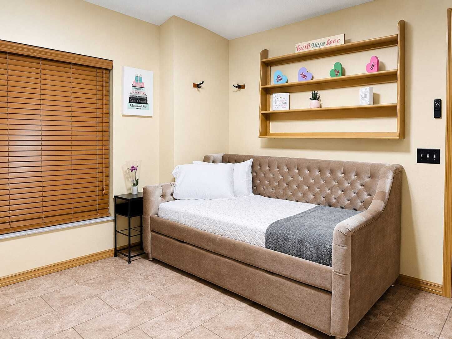 A bedroom with a beige sofa bed, a small bedside table with a vase, a wall-mounted shelf with decorative items, and a window with blinds.