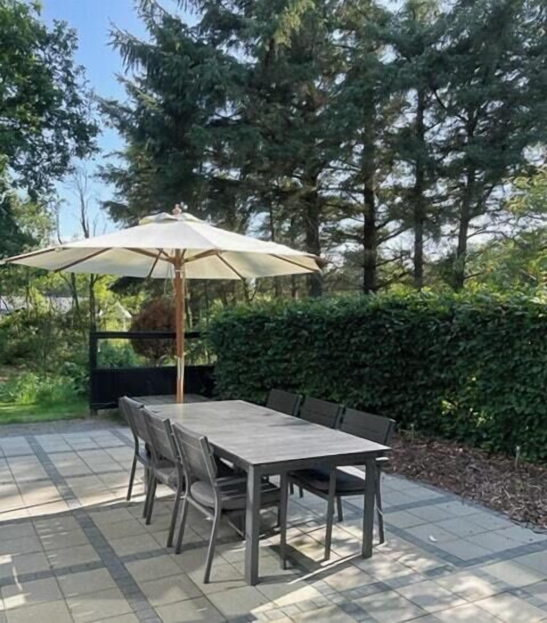 En udendørs terrasse med et bord, stole, en parasol og en hæk i baggrunden.