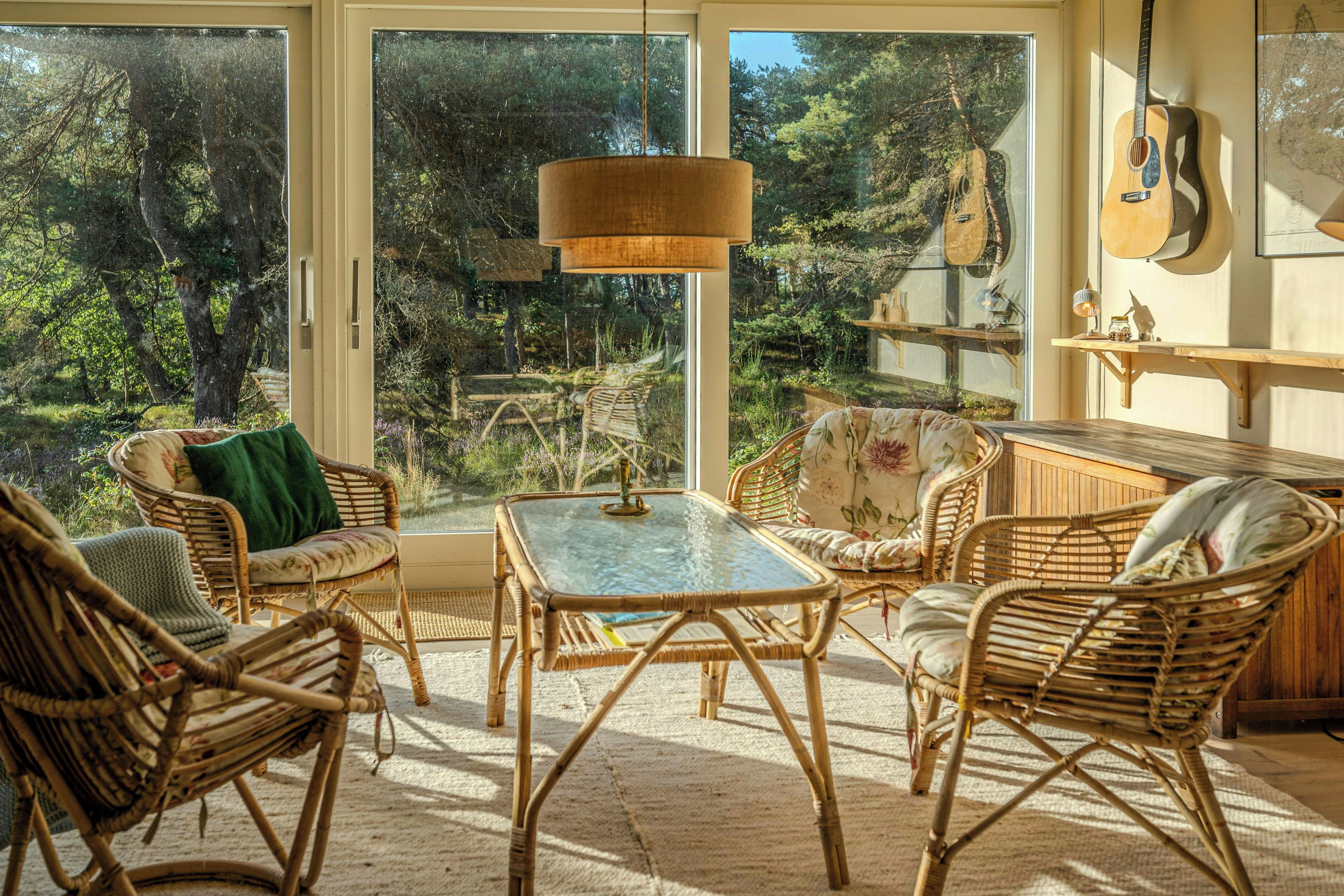 Quarto com móveis de vime, uma mesa de vidro e vista para uma floresta através de grandes janelas.