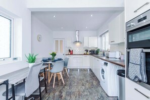 A modern kitchen with a built-in washing machine, a dining area with a table and chairs, and a clock on the wall. - Spacious 4BR Sleeps 7 | Contractor Stays (Hawarden)