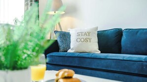 Ein blaues Sofa mit einem Kissen mit dem Aufdruck 'STAY COSY', ein Glas Orangensaft und ein Teller mit Croissants.