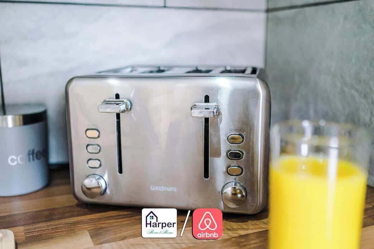 A stainless steel toaster on a kitchen counter with a glass of orange juice, a coffee mug, and logos for Harper Home & Home and Airbnb.