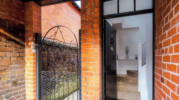 Fachada de ladrillo con una puerta de metal, una escalera que lleva al interior y un espacio interior blanco visible a través de la puerta.