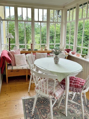 A conservatory with a round table, white chairs, a bench, and a view of greenery.