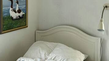 A bed with white bedding and a blue and white striped pillow, a white headboard, a wall-mounted lamp, and a framed picture of two ducks.