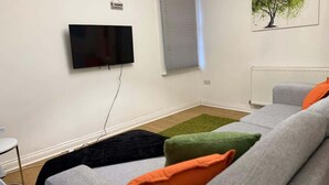 Ein Wohnzimmer mit Flachbildfernseher an der Wand, einem Fenster mit Jalousien, einer Heizung und einem Sofa mit bunten Kissen.