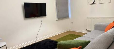 A living room with a flat-screen TV mounted on the wall, a window with blinds, a radiator, and a sofa with colorful cushions.