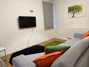 Ein Wohnzimmer mit Flachbildfernseher an der Wand, einem Fenster mit Jalousien, einer Heizung und einem Sofa mit bunten Kissen.
