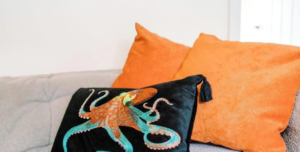 A bed with a grey blanket, an orange pillow, and a black pillow with an octopus design.