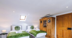 Et soveværelse med to senge, et træskab, et lille bord med en lampe og et ovenlysvindue.