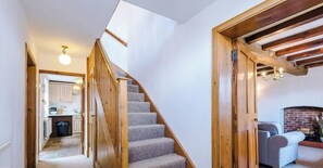 Een trap die leidt naar een leefruimte met een open haard, een keuken op de achtergrond, en een houten plafond.