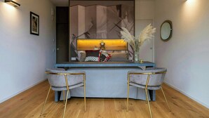 Una moderna recepción con encimera azul, dos sillones tapizados grises con patas doradas, un jarrón decorativo con plumas de miscanthus y un espejo montado en la pared.