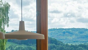 A dining area with a table and chairs, a hanging light fixture, and a view of a lake and forest through large windows. - Designer villa in Laven near Himmelbjerget (Silkeborg)