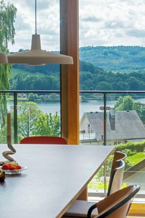 Ein Essbereich mit Tisch und Stühlen, einer Hängelampe und Blick auf einen See und Wald durch große Fenster.