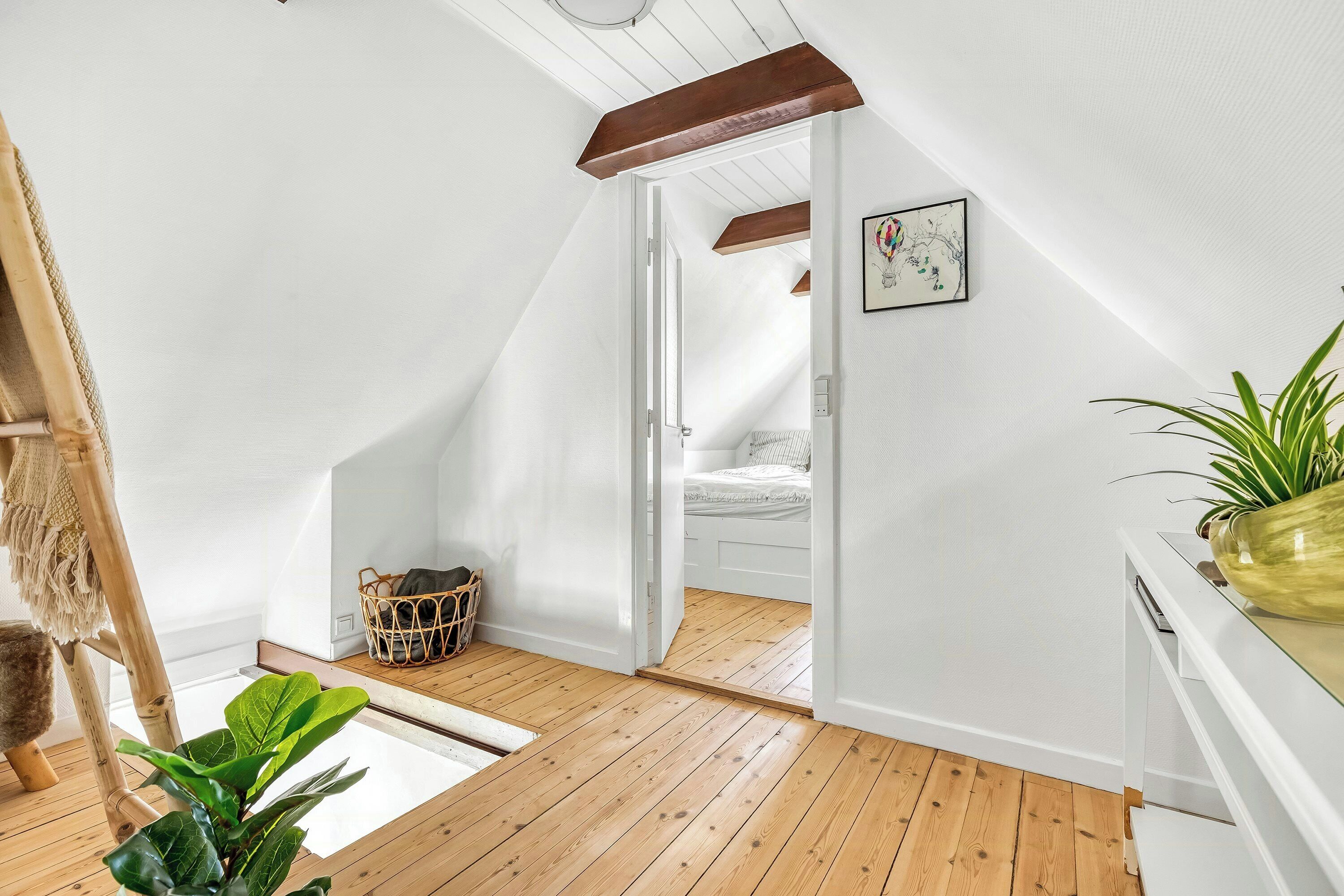 A loft bedroom with a sloped ceiling, wooden flooring, a bed, a small window, and a plant.