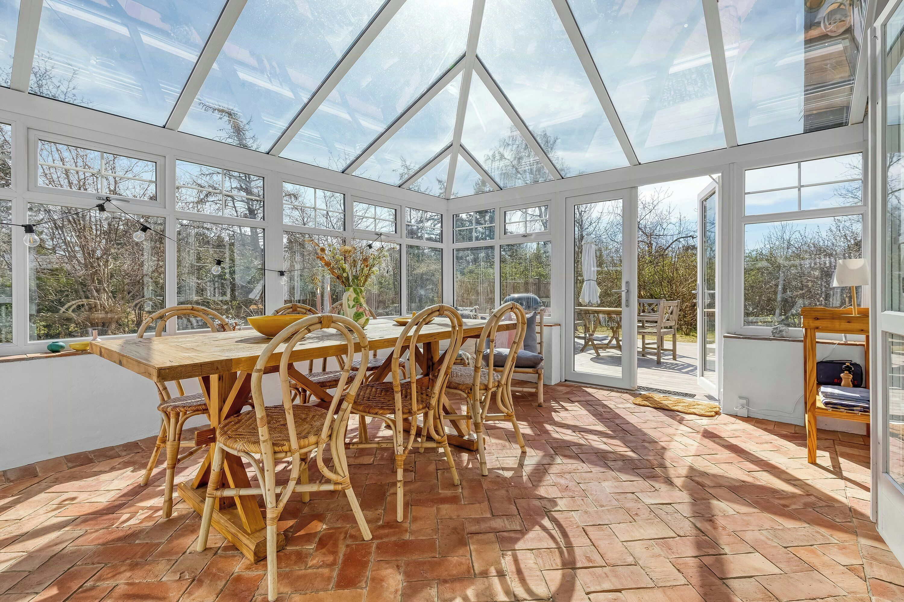 A conservatory with a wooden dining table and chairs, large windows, and a brick floor.