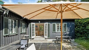 An outdoor patio with wooden benches and tables, a large umbrella, and a building with open shutters. - Summer house with large nature plot in Rørvig (Rørvig)