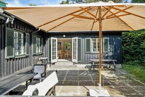 En uteplats med träbänkar och bord, ett stort parasoll och en byggnad med öppna fönsterluckor.