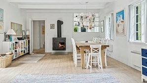 A cozy living room with a fireplace, a dining area with white chairs, and a bookshelf filled with books. - Summer house with large nature plot in Rørvig (Rørvig)