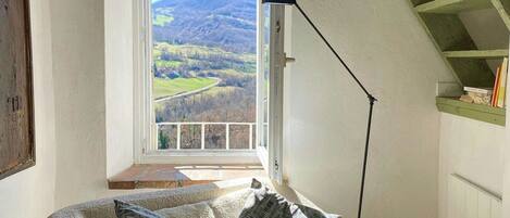 A cozy room with a sofa, pillows, a window with a view of the countryside, and a staircase.