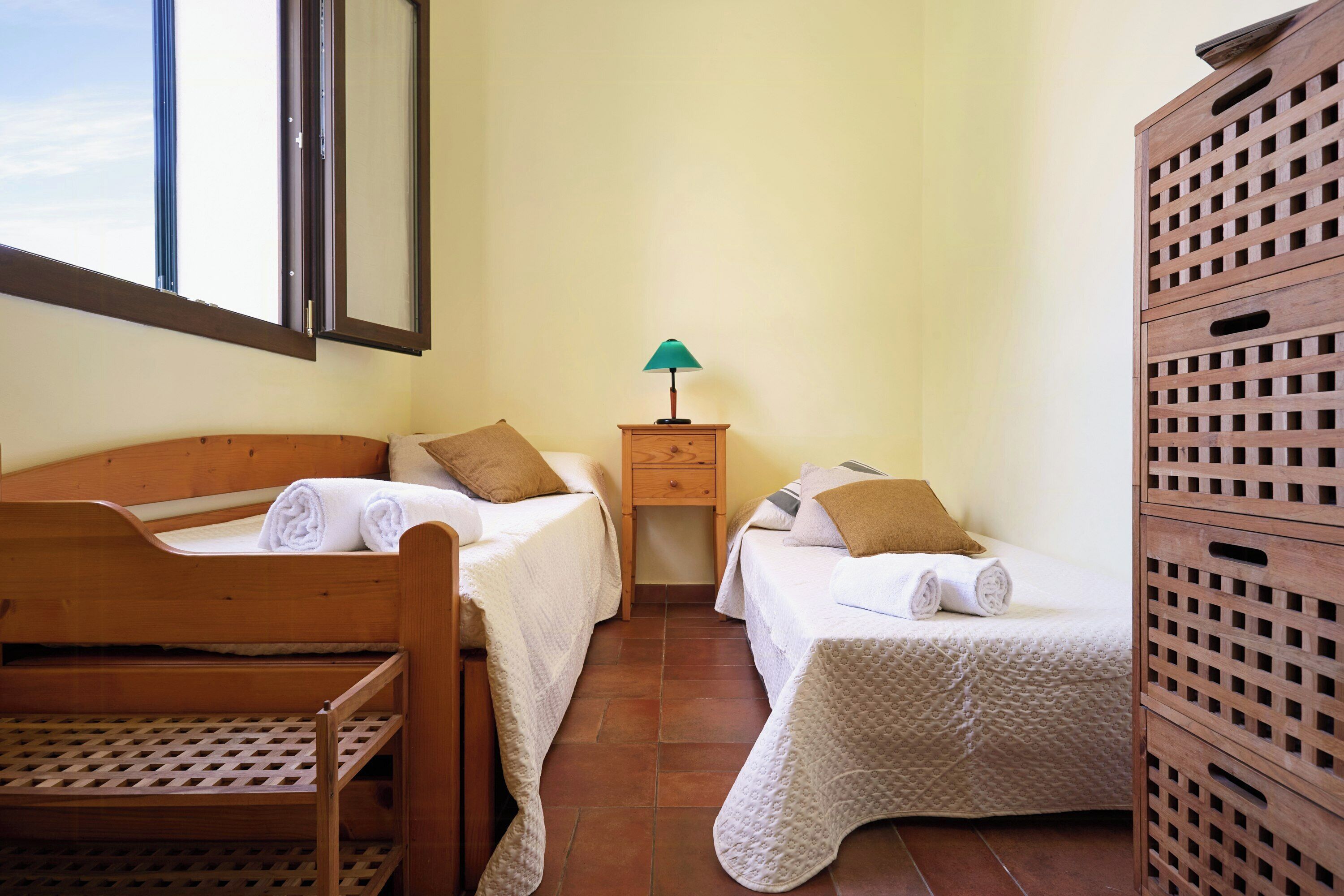 A bedroom with two beds, a nightstand with a lamp, a wooden wardrobe, and a window.