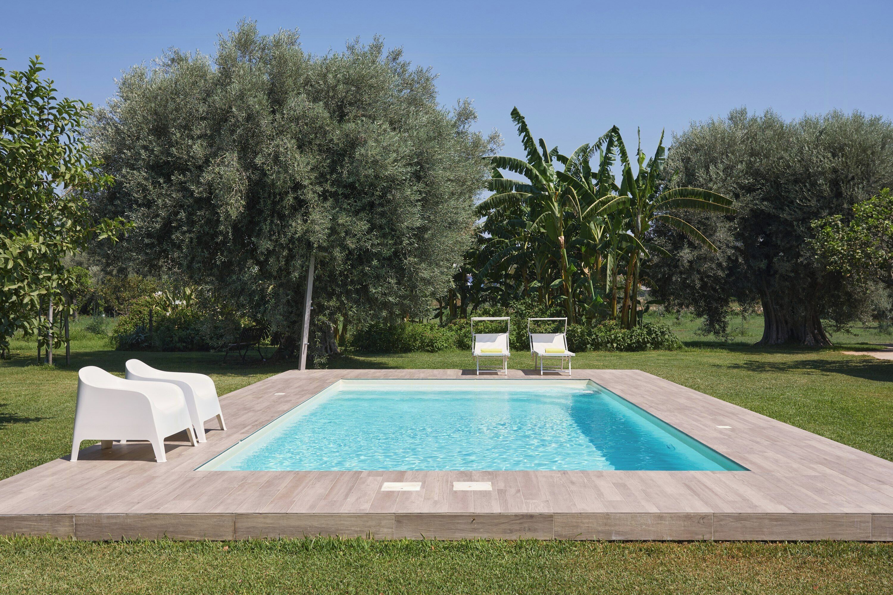 Une piscine rectangulaire, avec deux chaises longues blanches d’un côté et un décor arboré en arrière-plan.