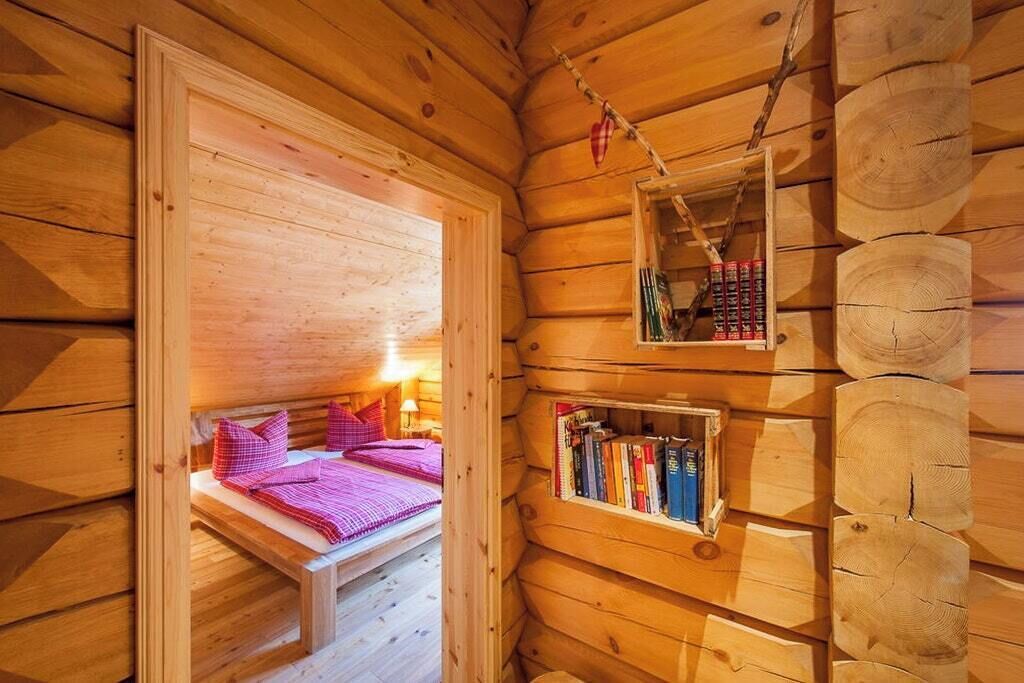 Ett mysigt rum i en stuga med en säng, en liten bokhylla och en hylla som hänger på väggen, fylld med böcker.