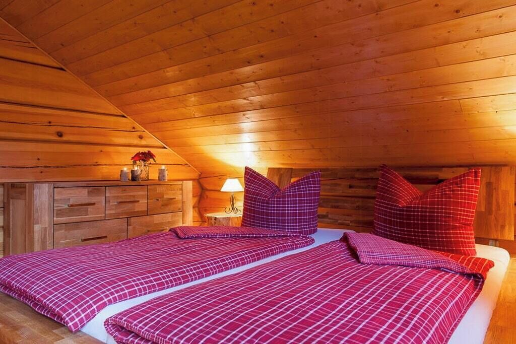 Un dormitori de cabana de fusta amb sostre inclinat, dos llits amb roba de llit vermella a quadradets, una nitzeta amb una làmpada i un armari de fusta.