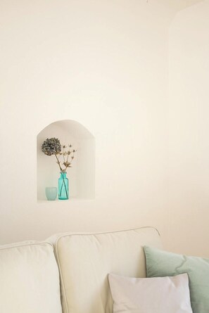 Ein Zimmer mit einer weißen Wand, einem kleinen Rundbogenfenster, einer türkisfarbenen Vase mit getrockneten Blumen und einem beigefarbenen Sofa mit Kissen.