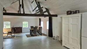Ein geräumiger Raum mit hoher Decke, freiliegenden Balken, einer Essecke mit Stühlen und Tisch, einem weißen Schrank und Fenstern, die für natürliches Licht sorgen.