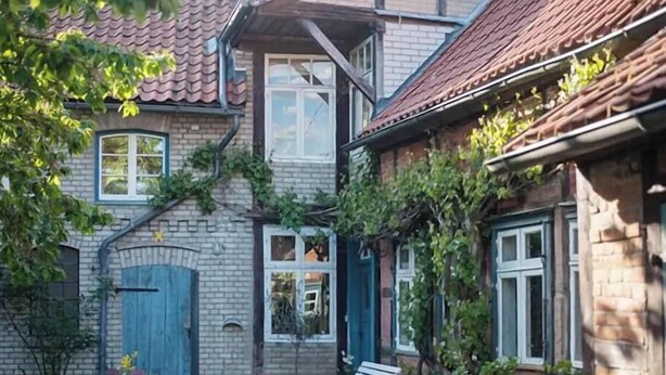 Et murstenshus med tegltag, en blå dør og en lille have med et bord, stole og en bænk.
