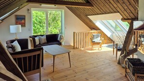 Une pièce de style « loft », avec un plancher en bois, un canapé, un fauteuil et une table. Une grande fenêtre offre une vue sur la verdure environnante.