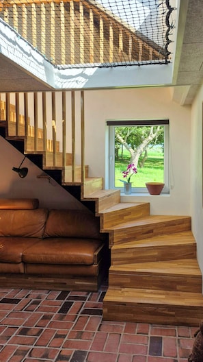 Un escalier en bois menant à un balcon, un canapé en cuir, une fenêtre donnant sur un paysage verdoyant et une plante en pot.