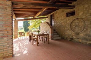 A covered patio with a stone wall, a wooden table with chairs, and a view of the outdoors. - Villa in the farmhouse with pool in Tuscany (Bucine)