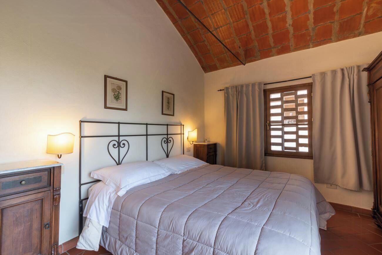 A bedroom with a bed, a nightstand, a dresser, a window with curtains, and a brick ceiling.