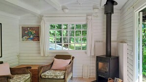 A cozy room with a wooden floor, a wicker chair, a bench, a fireplace, and a window with a view of greenery. - Retro and simplicity in two small houses by Arresø (Frederiksværk)