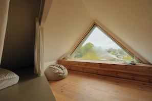 Ein Schlafzimmer mit schrägem Dach, einem Fenster mit Blick auf Grünflächen, Holzboden und einem Sitzsack.